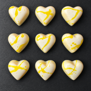 Nou bombons artesans en forma de cor de xocolata blanca, mel i llimona, amb decoració groga sobre fons antracita.